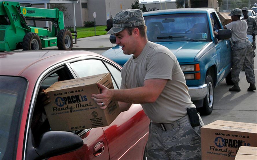 A man in military uniform handing a box labeled "Ready Meal" to a person in a car.