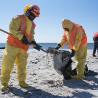 Two response workers in yellow and orange gear picking up a black substance with a shovel.