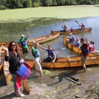 People sitting in canoes and standing on a shoreline.